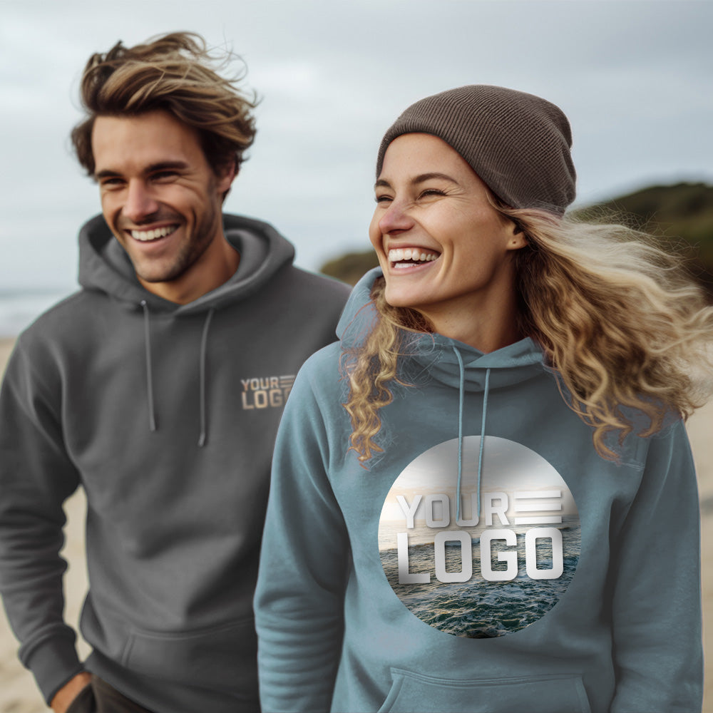 Lifestyle image of a man and a woman on the beach. The man is slightly behind the woman, wearing a rock colored sweatshirt with "your logo" on the left pocket area. The girl wears 