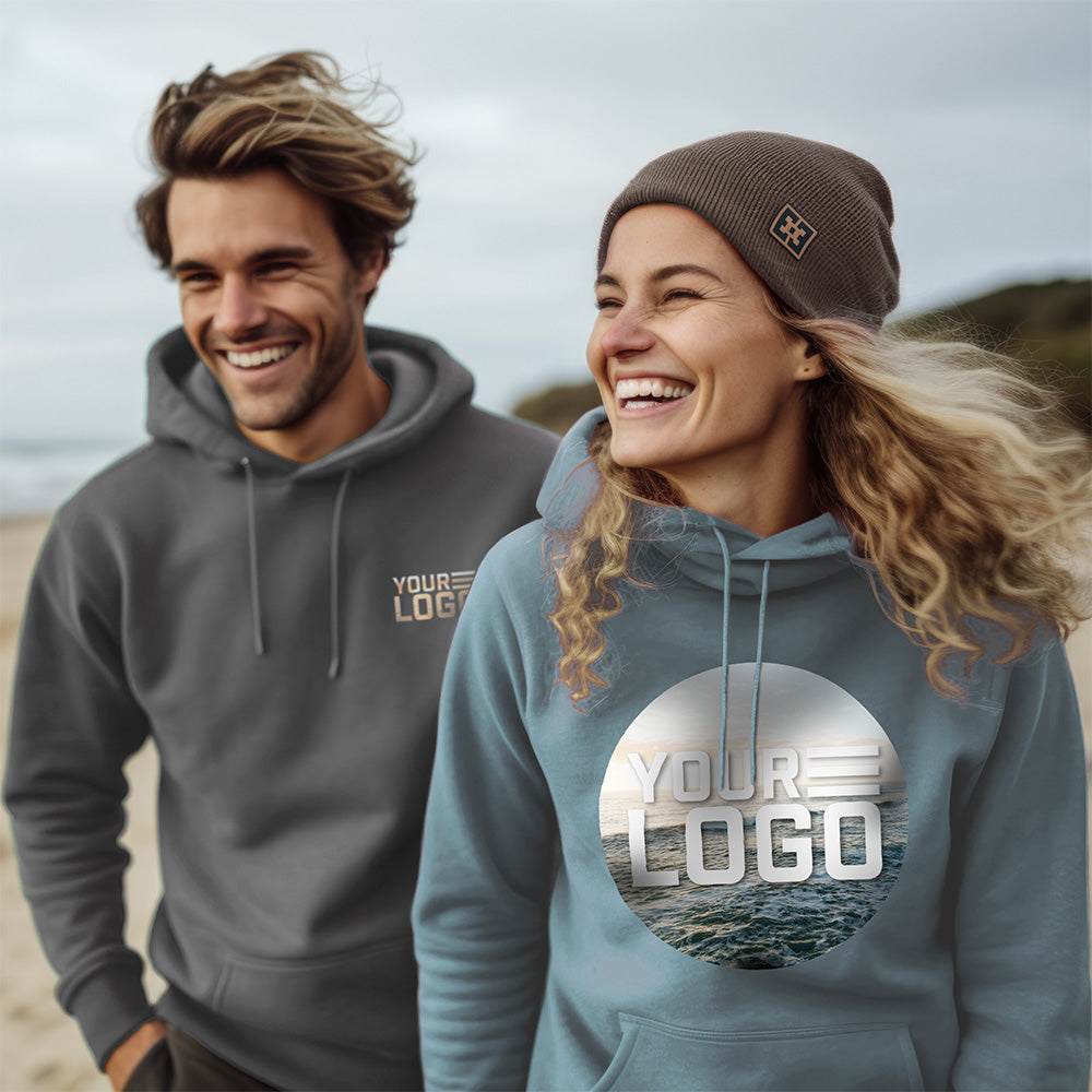 Lifestyle image of a man and a woman on the beach. The man stands slightly behind the woman, wearing a rock colored sweatshirt with "your logo" on the left pocket area. The woman wears a sage colored sweatshirt with a large "your logo" design on the front and a brown beanie with a Free Sunshields leather patch on the side. 