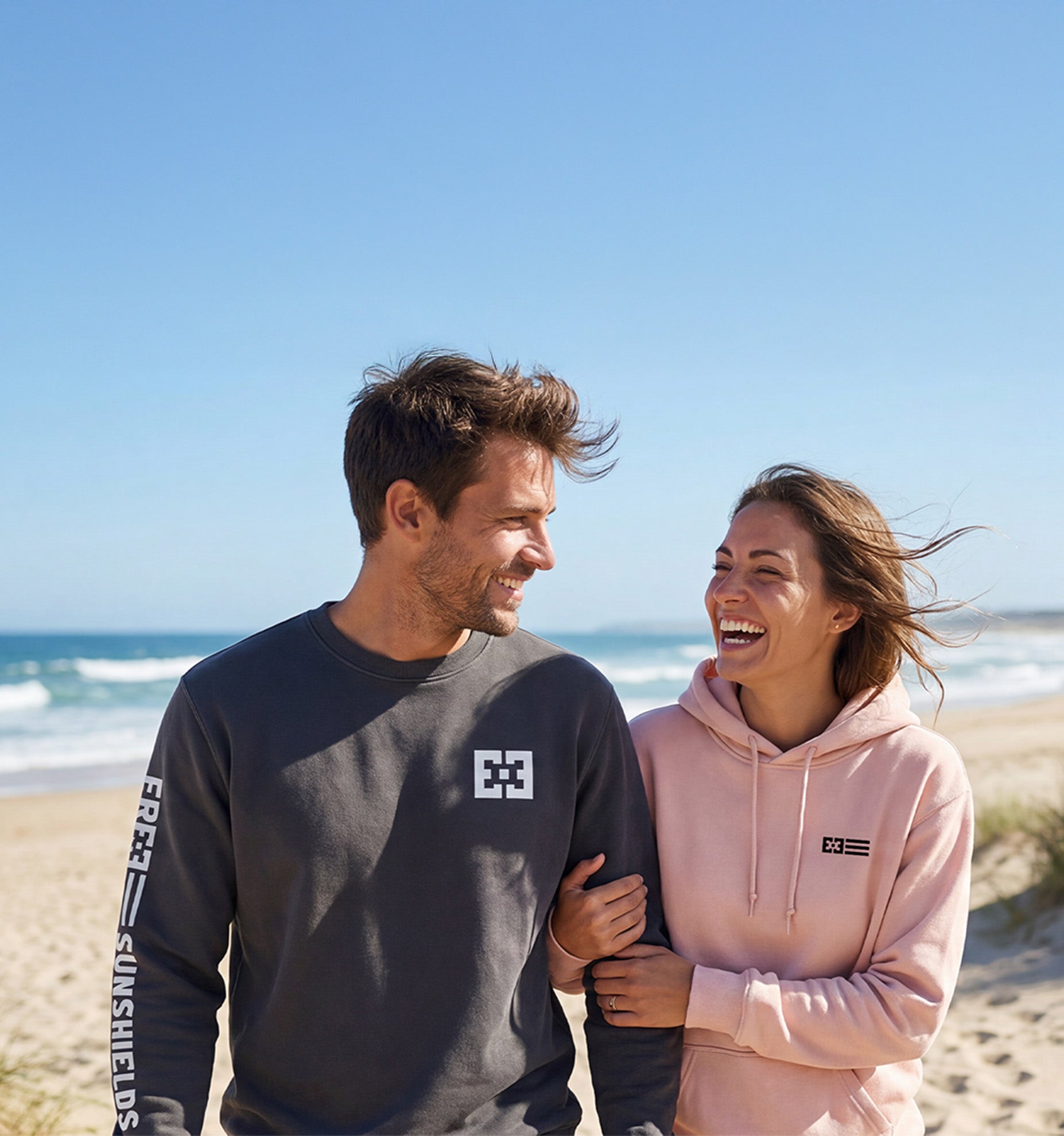 Couple walking and laughing on the beach wearing custom sweatshirts.